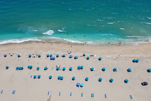 Aerial view of Singer Island beach featuring turquoise ocean waves sandy shore and rows of blue beach umbrellas with people in Riviera Beach Florida United States. Aerial view of Singer Island beach featuring turquoise ocean waves sandy shore and rows of blue beach umbrellas with people in Riviera Beach Florida United States.