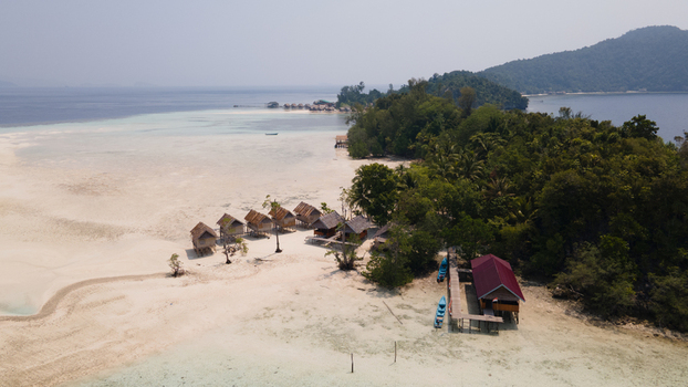 Aerial view of Ransiwor homestay featuring wooden huts on a white sand beach surrounded by turquoise water and lush tropical vegetation in Raja Ampat Southwest Papua Indonesia.