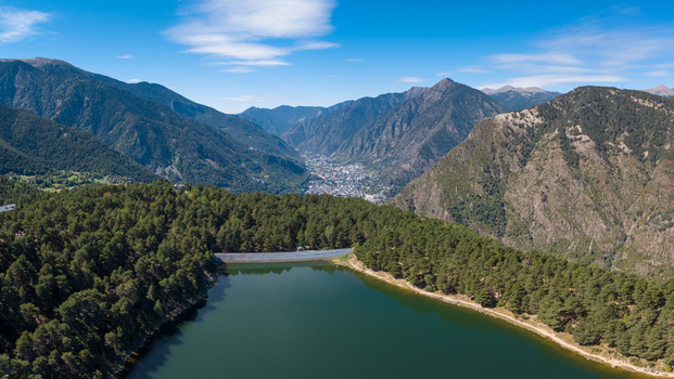 Aerial view of Lake Engolasters surrounded by dense pine forests with the mountains of the Pyrenees and the town of Andorra La Vella in the distance Encamp Andorra.
