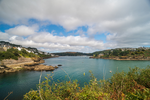 View of the Fowey River estuary with the town of Fowey on the hillside featuring historic buildings small boats and rocky shores in Fowey Cornwall United Kingdom. View of the Fowey River estuary with the town of Fowey on the hillside featuring historic buildings small boats and rocky shores in Fowey Cornwall United Kingdom.