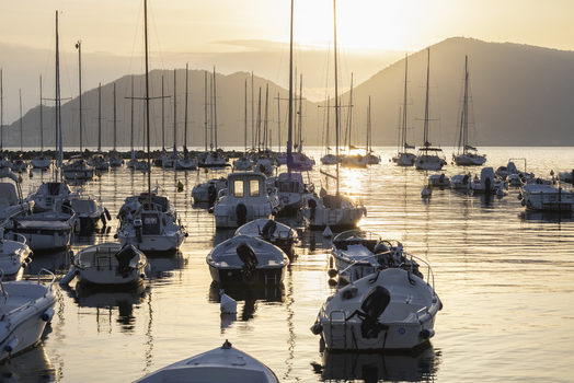  Lerici Italy 08 March 2026 View of a harbor filled with boats their masts reaching towards the sky as the sun sets behind the distant mountains painting the water in warm golden hues.