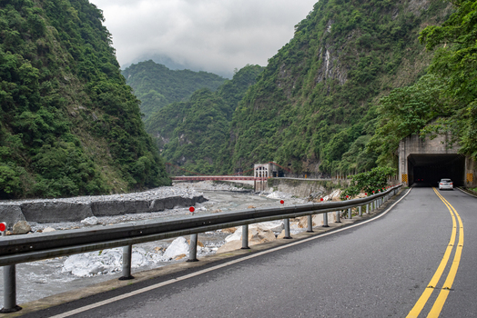 View of a winding road leading into a tunnel in the dramatic mountain gorge of Taroko National Park with a rocky riverbed and lush green slopes in Xiulin Township Hualien County Taiwan.