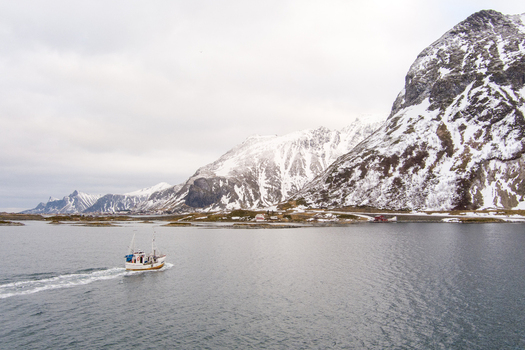 Aerial view of a small fishing boat navigating the calm waters of a bay in Lofoten surrounded by dramatic snow capped mountains under a cloudy sky in Nordland Norway.