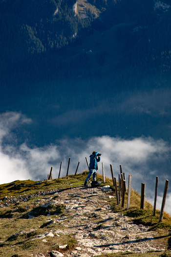 View of a photographer on a mountain ridge in the Swiss Alps capturing the landscape with a camera with a wooden fence and clouds drifting below in Airolo Ticino Switzerland. View of a photographer on a mountain ridge in the Swiss Alps capturing the landscape with a camera with a wooden fence and clouds drifting below in Airolo Ticino Switzerland.