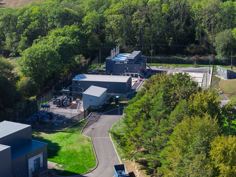 Aerial view of a data center facility with industrial buildings cooling units and fenced perimeter surrounded by lush green trees in the United Kingdom.