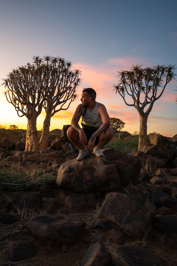 Aussenkehr Namibia 06 March 2026 View of a lone figure perched atop a dark rock framed by quiver trees against a pastel sky creating a serene yet striking contrast. Aussenkehr Namibia 06 March 2026 View of a lone figure perched atop a dark rock framed by quiver trees against a pastel sky creating a serene yet striking contrast.