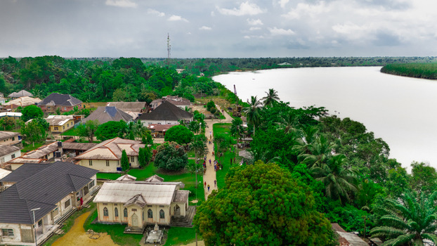 Aerial view of a riverfront settlement with houses lush green trees and a wide river under a cloudy sky in Rivers Nigeria.