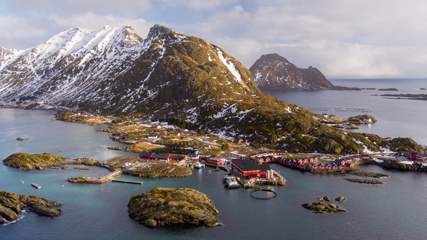 Aerial view of the Lofoten archipelago featuring traditional red wooden fishing cabins snow capped mountains and fish farming pens in the clear coastal waters of Nordland Norway.