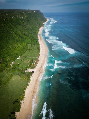 Aerial view of Pantai Nunggalan with sandy beach turquoise waves crashing against the shore and lush green cliffs rising above the coastline in Kecamatan Kuta Selatan Bali Indonesia.
