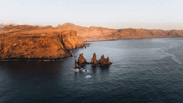 Aerial view of basalt sea stacks rising sharply from the dark ocean waters framed by rugged cliffs under the soft glow of dawn Reynisdrangar Iceland.
