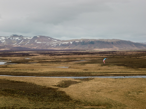 Aerial view of a kitesurfer gliding over a river in a vast Icelandic landscape with snow capped mountains in the distance in Borgarbyggd Iceland. Aerial view of a kitesurfer gliding over a river in a vast Icelandic landscape with snow capped mountains in the distance in Borgarbyggd Iceland.