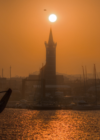 View of the church tower silhouetted against the bright sunset over the harbor with boats moored in the water in Civitanova Marche Marche Italy. View of the church tower silhouetted against the bright sunset over the harbor with boats moored in the water in Civitanova Marche Marche Italy.