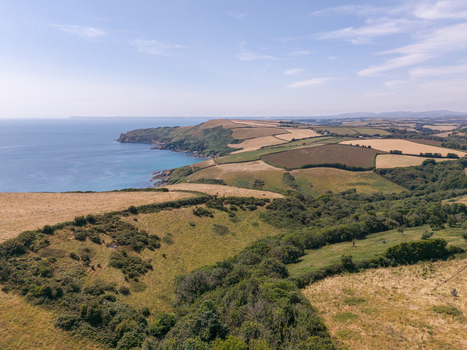 Aerial view of Lansallos beach and the surrounding rugged coastline featuring rolling green fields and cliffs meeting the sea under a clear blue sky in Lansallos England United Kingdom. Aerial view of Lansallos beach and the surrounding rugged coastline featuring rolling green fields and cliffs meeting the sea under a clear blue sky in Lansallos England United Kingdom.