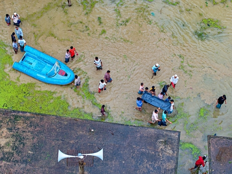 Banshkhali Upazila Chittagong Division Bangladesh 10 July 2026 Aerial view of people wading through murky floodwaters and carrying a stretcher near a blue inflatable boat. Banshkhali Upazila Chittagong Division Bangladesh 10 July 2026 Aerial view of people wading through murky floodwaters and carrying a stretcher near a blue inflatable boat.