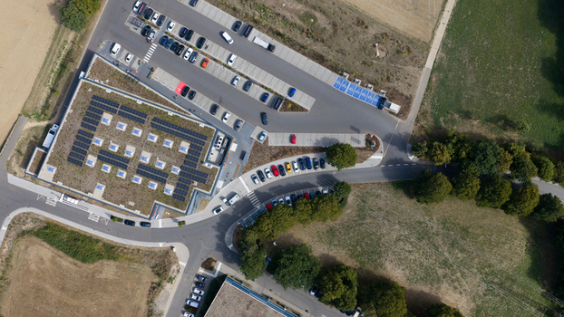 Aerial view of a building with a green roof solar panels and skylights adjacent to a parking lot with cars surrounded by trees and fields in Liederbach am Taunus Hessen Germany.