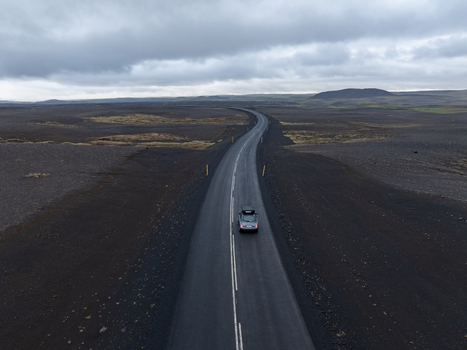 Aerial view of a car driving along a winding asphalt road through a vast desolate volcanic landscape with dark lava fields under a cloudy sky in Iceland. Aerial view of a car driving along a winding asphalt road through a vast desolate volcanic landscape with dark lava fields under a cloudy sky in Iceland.