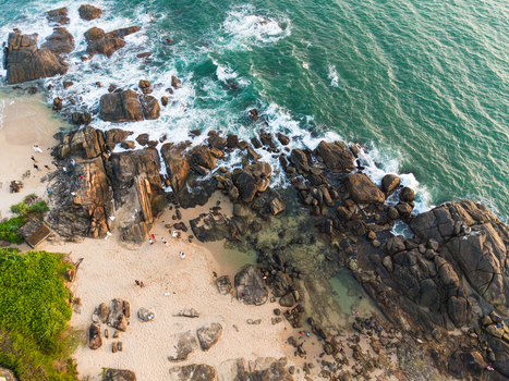 Aerial view of rocky coastline with turquoise waves crashing against dark boulders and people walking on the sandy beach Galle Southern Province Sri Lanka. Aerial view of rocky coastline with turquoise waves crashing against dark boulders and people walking on the sandy beach Galle Southern Province Sri Lanka.