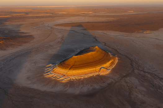 Aerial view of a solitary plateau with layered rock formations illuminated by the setting sun casting a long shadow over the arid desert landscape Mangystau Region Kazakhstan. Aerial view of a solitary plateau with layered rock formations illuminated by the setting sun casting a long shadow over the arid desert landscape Mangystau Region Kazakhstan.