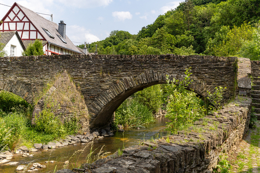 View of a historic stone arch bridge over the Elzbach river with a traditional half timbered house and lush green forest in Monreal Rhineland Palatinate Germany. View of a historic stone arch bridge over the Elzbach river with a traditional half timbered house and lush green forest in Monreal Rhineland Palatinate Germany.