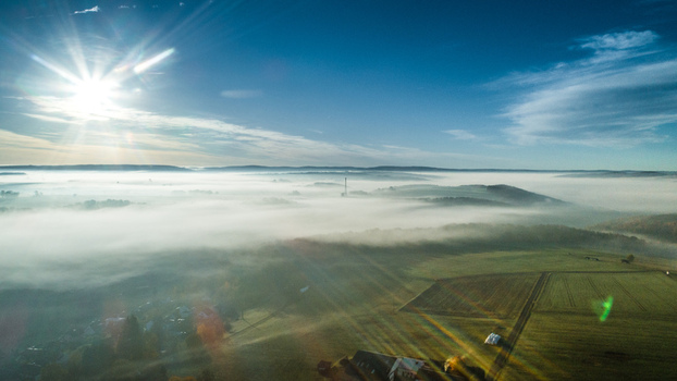 Aerial view of a rural landscape covered in a thick layer of morning fog with sun rays breaking through the sky and illuminating the fields and trees in Germany. Aerial view of a rural landscape covered in a thick layer of morning fog with sun rays breaking through the sky and illuminating the fields and trees in Germany.