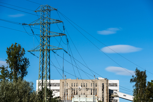 View of a green metal electricity transmission tower with power lines against a clear blue sky and an industrial building in the background in Poland.