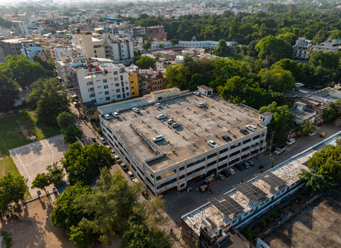 Aerial view of a multi level parking structure in Udaipur Rajasthan India surrounded by urban buildings lush green trees and a basketball court.