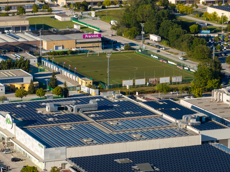 Aerial view of a large commercial building with extensive solar panel arrays on the roof adjacent to a green soccer field and paved parking areas in Ancona Marche Italy. Aerial view of a large commercial building with extensive solar panel arrays on the roof adjacent to a green soccer field and paved parking areas in Ancona Marche Italy.
