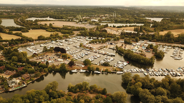 Aerial view of the Thames River marina filled with numerous boats docked along the banks surrounded by lush green trees and parkland in the United Kingdom. Aerial view of the Thames River marina filled with numerous boats docked along the banks surrounded by lush green trees and parkland in the United Kingdom.