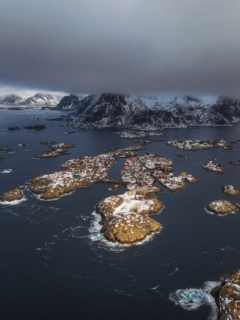 Aerial view of the fishing village nestled on rocky islands amidst the dark sea a stark contrast to the snow capped mountains under a brooding sky Henningsv&aelig;r Nordland Norway.