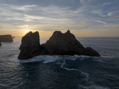 Aerial view of rocky sea stacks rising from the ocean with waves crashing against the base during a golden sunset in Nueva Asturias Spain.