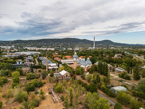 Aerial view of the historic Obuda Gasworks with brick industrial buildings towers and chimneys amid trees urban development and the Buda Hills in Budapest Hungary. Aerial view of the historic Obuda Gasworks with brick industrial buildings towers and chimneys amid trees urban development and the Buda Hills in Budapest Hungary.