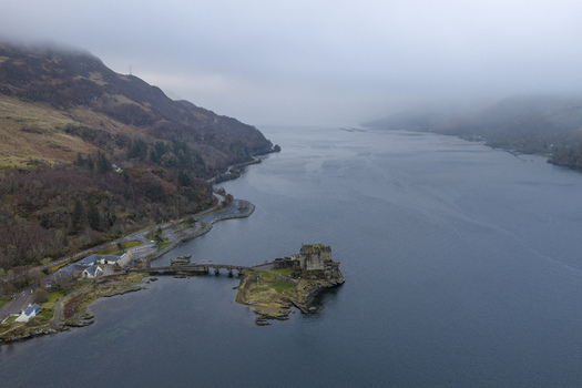 Aerial view of Eilean Donan Castle on its tidal island connected by a stone bridge to the shore of Loch Duich under a misty sky Dornie Scotland United Kingdom.