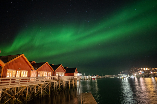 View of vibrant green aurora borealis dancing over traditional red wooden cabins on stilts along the waterfront in Leknes Lofoten Norway.