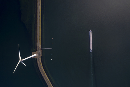 Aerial view of a wind turbine standing on a narrow causeway next to a cargo ship navigating the dark waters of the Krammer locks in Bruinisse Zeeland Netherlands. Aerial view of a wind turbine standing on a narrow causeway next to a cargo ship navigating the dark waters of the Krammer locks in Bruinisse Zeeland Netherlands.