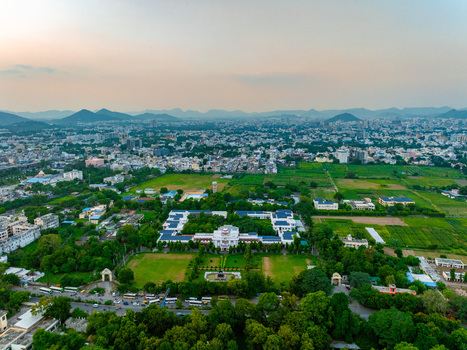 Aerial view of a large white building complex surrounded by lush green fields and trees in Udaipur Rajasthan India. Aerial view of a large white building complex surrounded by lush green fields and trees in Udaipur Rajasthan India.