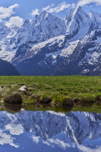 View of the Engadin valley with snow capped mountains reflecting in a calm pond and a green meadow in the foreground in Grisons Switzerland. View of the Engadin valley with snow capped mountains reflecting in a calm pond and a green meadow in the foreground in Grisons Switzerland.