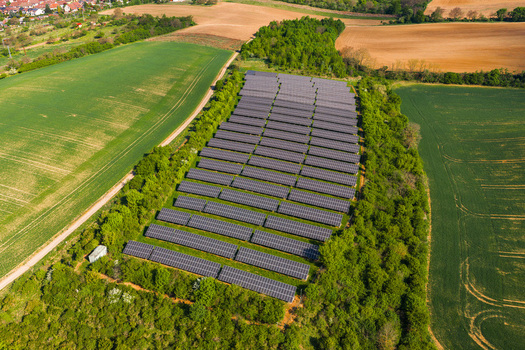 Aerial view of a solar farm with rows of photovoltaic panels surrounded by lush green fields trees and a dirt path in Kyjov South Moravian Region Czechia.