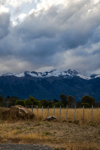 View of the snow capped Andes in Parque Nacional Los Alerces with a golden field wooden fence and large boulders under a dramatic cloudy sky in Futaleufu Department Chubut Province Argentina. View of the snow capped Andes in Parque Nacional Los Alerces with a golden field wooden fence and large boulders under a dramatic cloudy sky in Futaleufu Department Chubut Province Argentina.