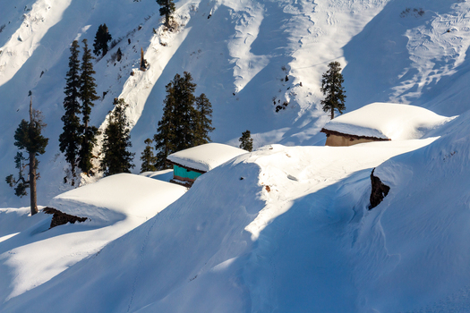 View of snow laden roofs of small buildings peek out from the mountainside contrasting with the dark green trees in Haveli Azad Kashmir Pakistan.