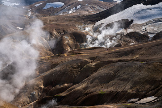 View of geothermal steam rises above the rugged terrain contrasting with the stark white snowfields clinging to the distant peaks Hrunamannahreppur Iceland.