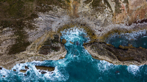 Aerial view of Lulworth Cove with turquoise waves crashing against rugged limestone cliffs and rocky coastline in the United Kingdom.