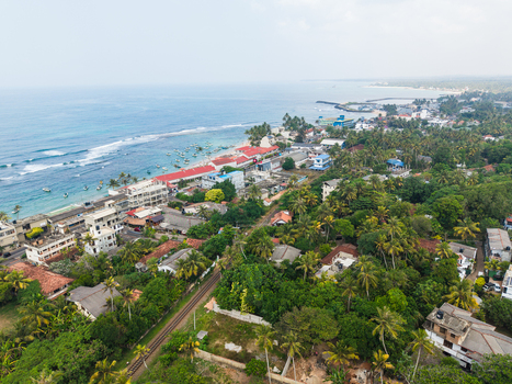 Aerial view of the coastal town of Hikkaduwa featuring tropical palm trees residential buildings and the turquoise Indian Ocean coastline in Hikkaduwa Southern Province Sri Lanka. Aerial view of the coastal town of Hikkaduwa featuring tropical palm trees residential buildings and the turquoise Indian Ocean coastline in Hikkaduwa Southern Province Sri Lanka.
