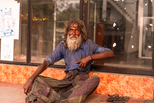 Sri Lanka 14 January 2026 View of an elderly man with long matted hair and a beard wearing a blue shirt and sarong while sitting in front of a glass storefront with posters.