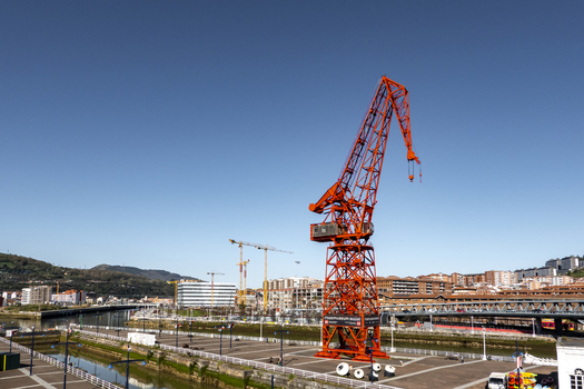 View of the iconic red Carola Crane standing on the waterfront of the Nervion River with city buildings and a clear blue sky Greater Bilbao Basque Country Spain. View of the iconic red Carola Crane standing on the waterfront of the Nervion River with city buildings and a clear blue sky Greater Bilbao Basque Country Spain.