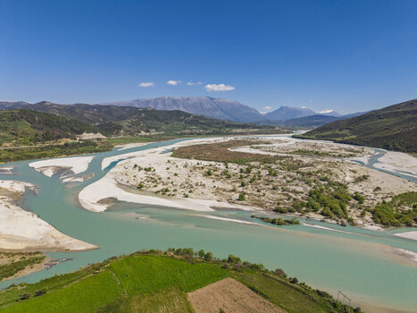 Aerial view of Vjosa Wild River National Park with its winding turquoise river expansive gravel riverbeds lush green fields and surrounding mountains in Permet Gjirokaster County Albania. Aerial view of Vjosa Wild River National Park with its winding turquoise river expansive gravel riverbeds lush green fields and surrounding mountains in Permet Gjirokaster County Albania.
