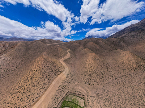 Aerial view of a winding dirt road cutting through arid mountainous terrain with sparse vegetation under a bright blue sky with scattered clouds in Nepal.