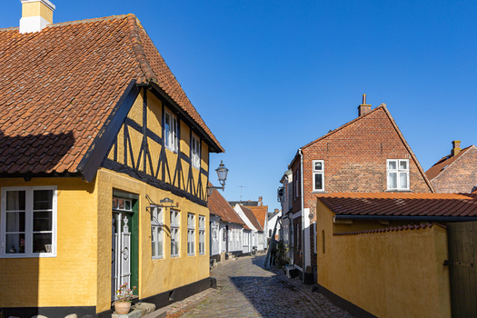 View of historic half timbered houses with yellow facades and red tiled roofs along a narrow cobblestone street under a clear blue sky in Ribe Denmark. View of historic half timbered houses with yellow facades and red tiled roofs along a narrow cobblestone street under a clear blue sky in Ribe Denmark.