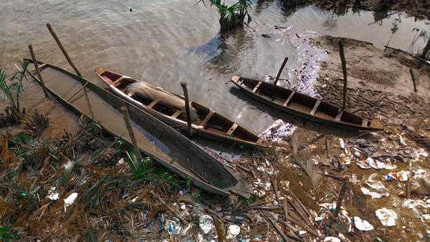 Aerial view of wooden dugout canoes moored along a muddy riverbank scattered with plastic debris and palm fronds in Minima Rivers Nigeria.
