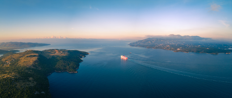 Aerial view of a large ferry cruising through a calm sea strait between lush green hills and islands under a soft sunset sky Ksamil Vlore County Albania. Aerial view of a large ferry cruising through a calm sea strait between lush green hills and islands under a soft sunset sky Ksamil Vlore County Albania.