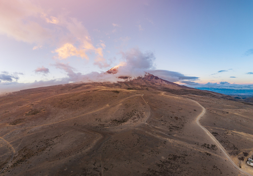 Aerial view of Volcano Chimborazo with its peak partially obscured by clouds featuring an arid landscape and a winding dirt road under a soft sunset sky in Guano Chimborazo Ecuador. Aerial view of Volcano Chimborazo with its peak partially obscured by clouds featuring an arid landscape and a winding dirt road under a soft sunset sky in Guano Chimborazo Ecuador.